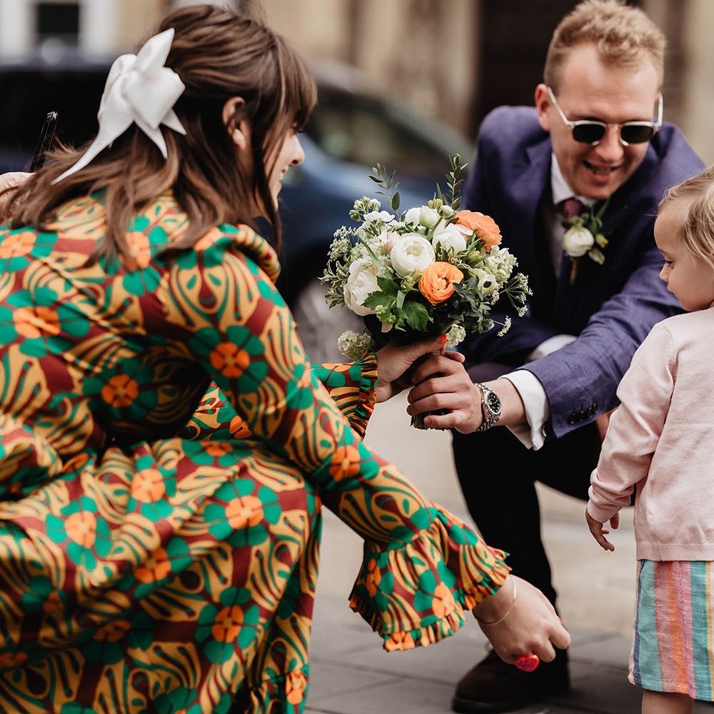 Bride wears brightly coloured dress complete with white bow in her hair holds white and peach coloured floral bouquet 