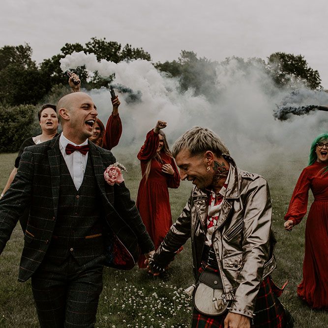 Groom walk through grey smoke bombs beside their bridesmaids who wear long sleeve gowns 