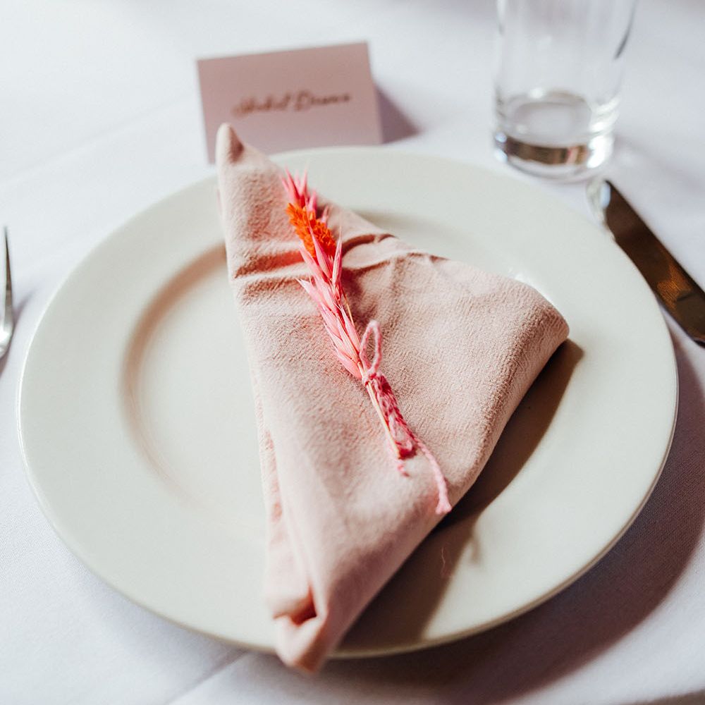 Pale pink fabric napkin lays across white plate finished with small coral dried floral decor tied with pink string