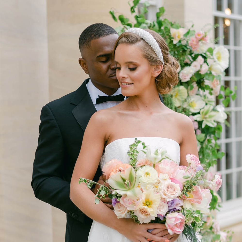 Groom in black tie embraces the bride from behind who wears a strapless wedding dress and beaded headband holding a colourful bouquet with coral pink nails 