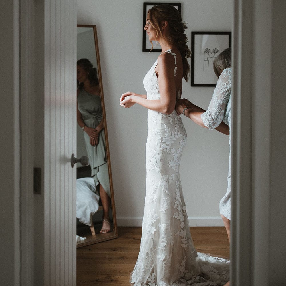 Bride wears floral embellished fitted Enzoani wedding dress and stands in front of the mirror on the morning of her wedding day