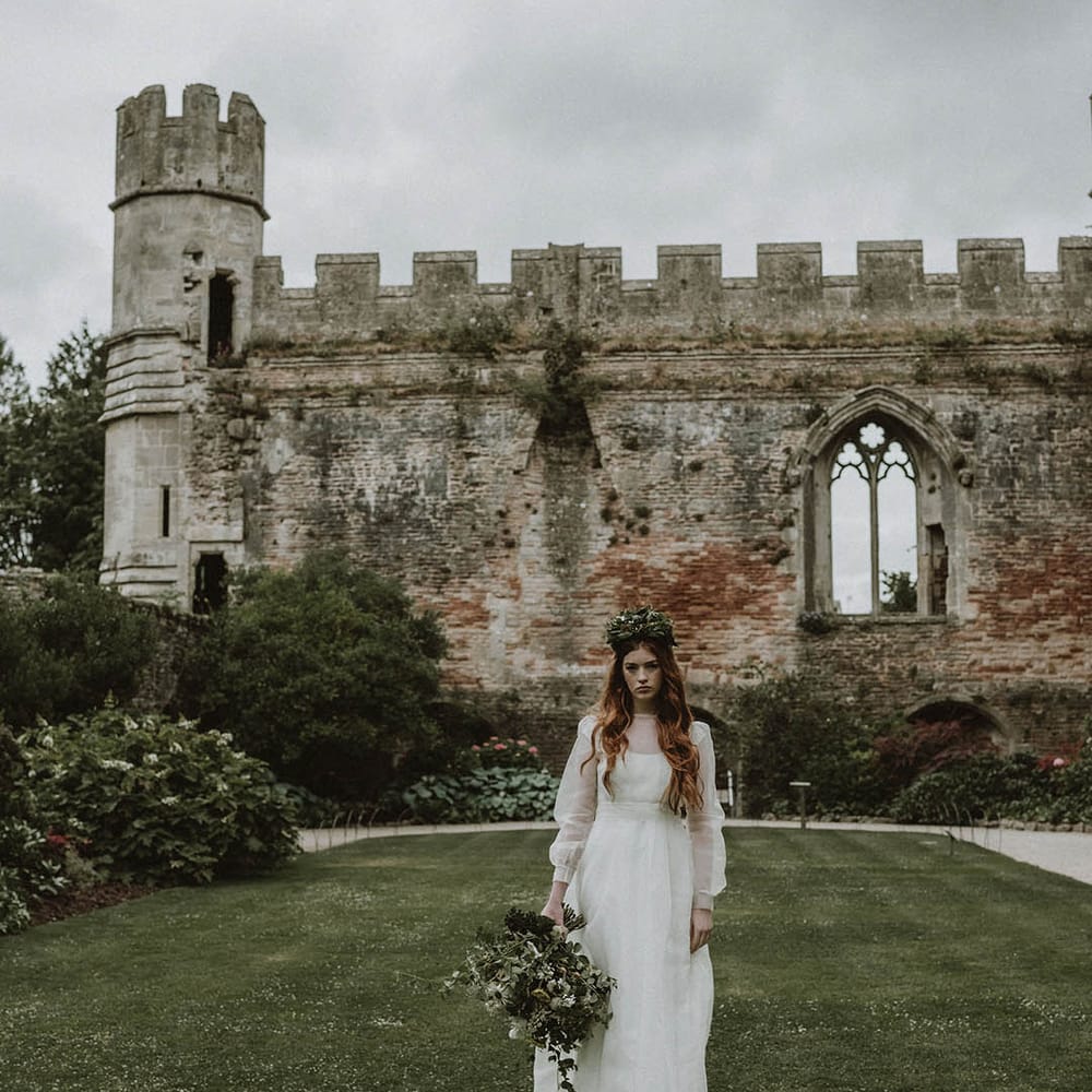 Bride in a wedding dress with sheer top layer and long sleeves wearing a foliage flower crown and holding a foliage bouquet standing in the gardens at Bishop's Palace, Wells