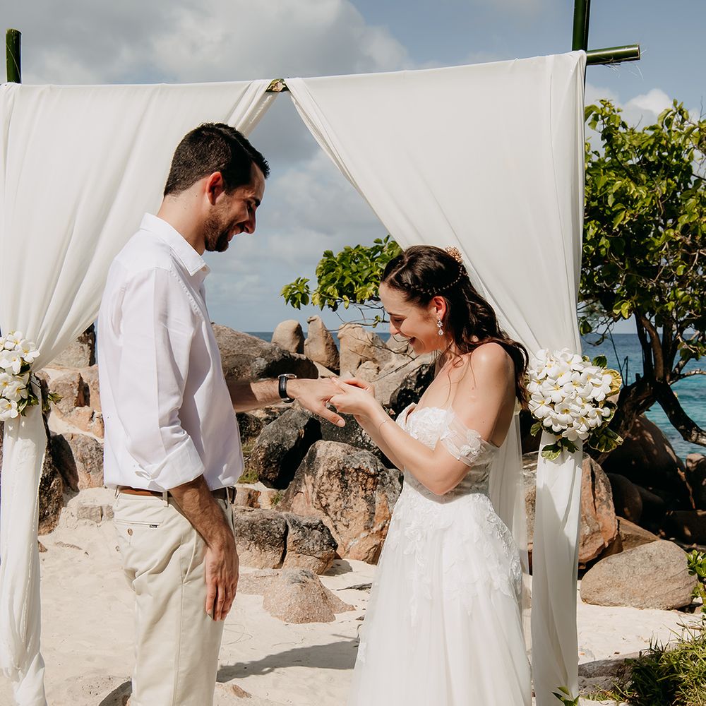 Bride in off shoulder lace wedding dress with sweetheart neckline and cathedral veil putting wedding ring on groom wearing white shirt and light coloured chinos