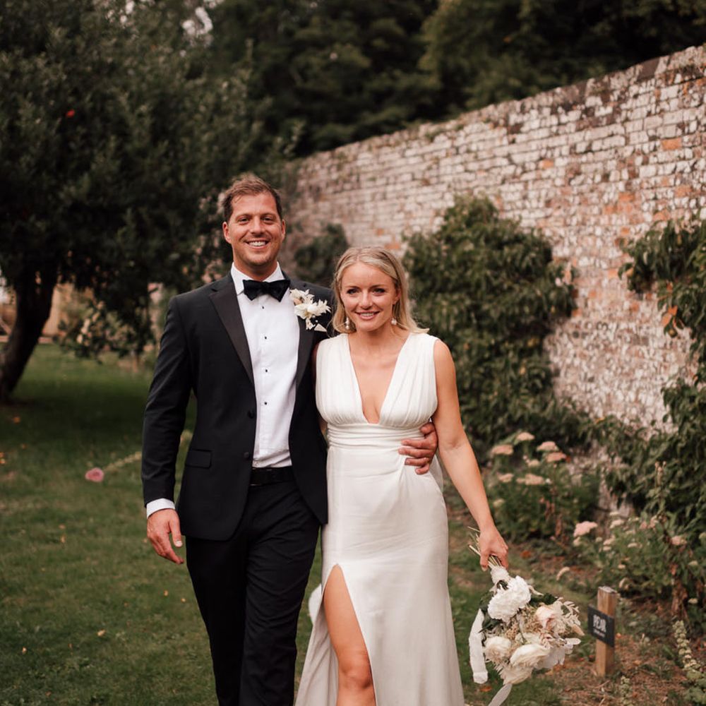 Groom in black tie with white flower buttonhole walking with the bride in a plunging satin wedding dress and orchid bouquet 