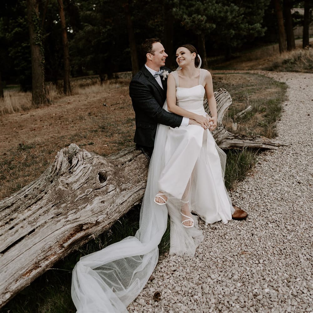 Groom in twee suit jacket sits on a tree with the bride on his lap wearing a sparkly wedding dress laughing together 