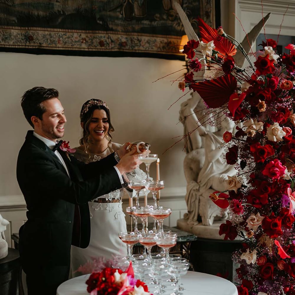 Groom in classic black tuxedo with magenta amaryllis and orchid boutonniere and bride in two piece bridal separates pouring pink champagne into their wedding champagne tour at Elmore Court
