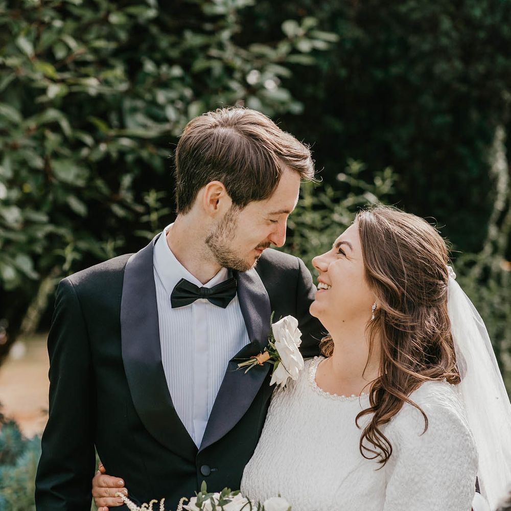 Bride in textured wedding dress and shoulder length veil smiles up at the groom in a black tuxedo with white rose buttonhole 