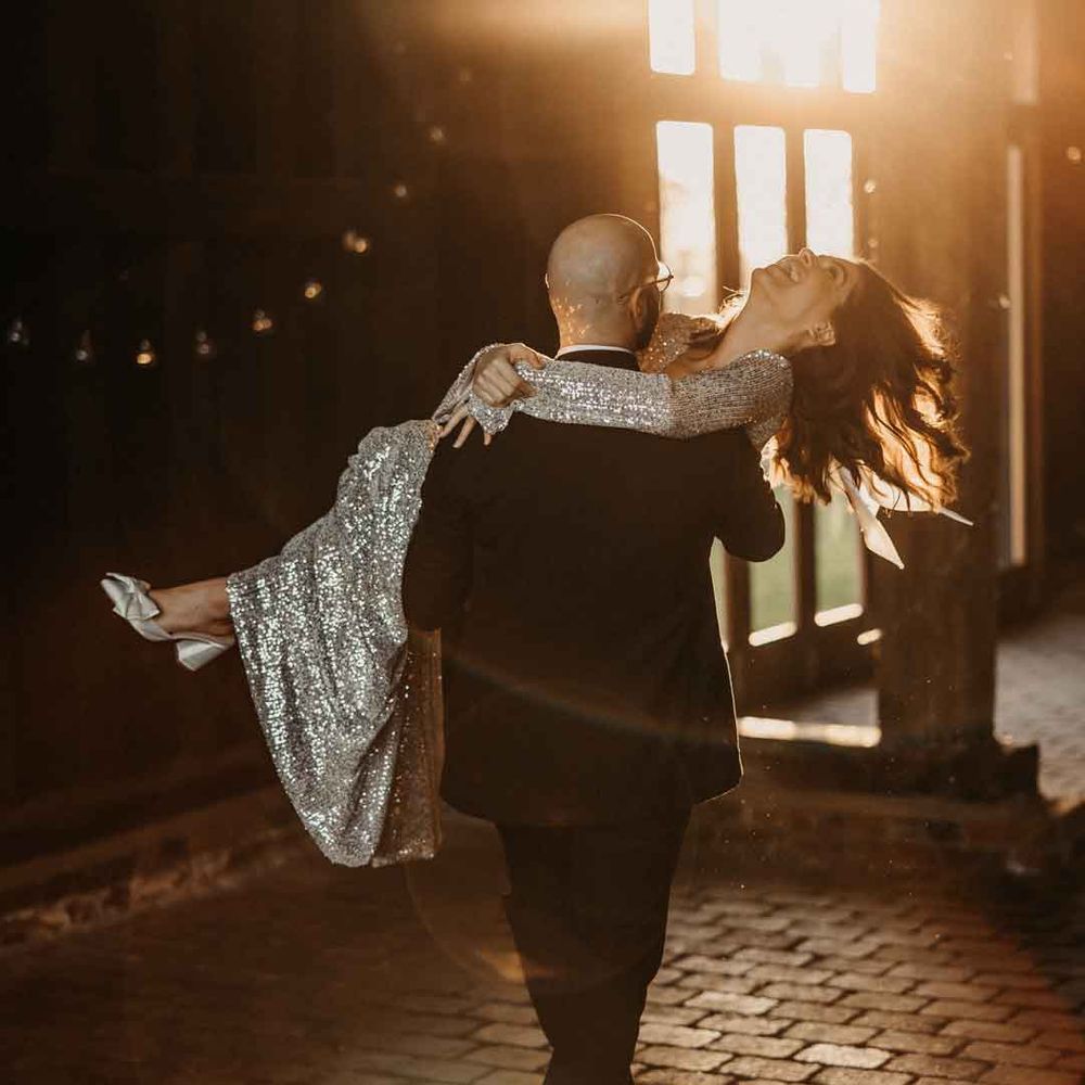 Bride in silver sparkly wedding dress and strapless wedding heels being carried by groom in classic black tuxedo