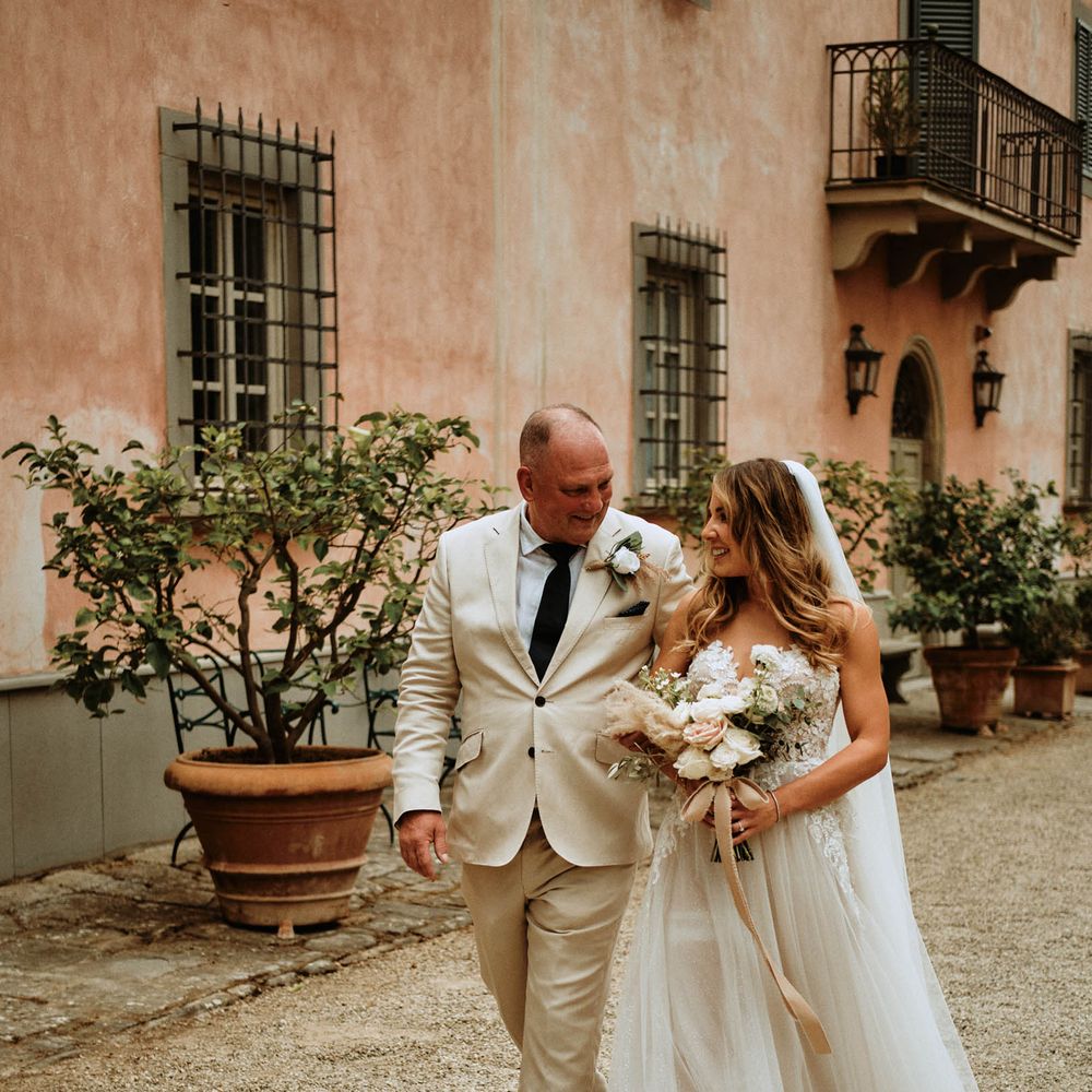Father of the bride in linen suit walking with bride in embroidered tulle wedding dress and white rose bridal bouquet at Italian destination wedding