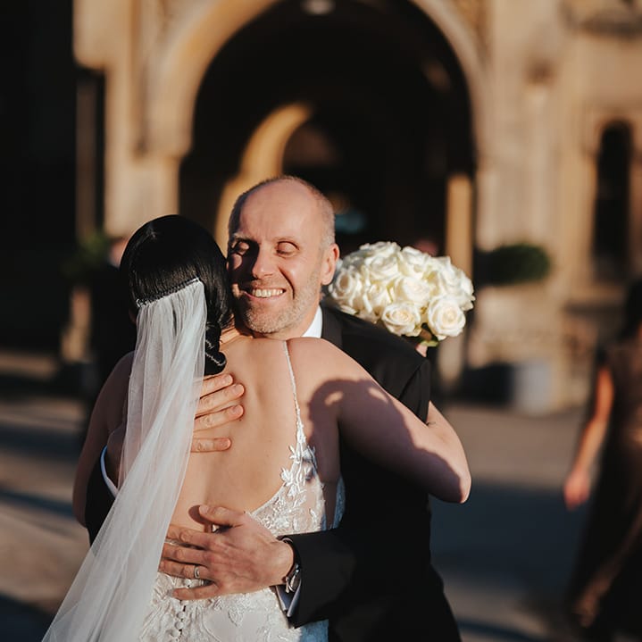 Bride in low scoop back lace wedding dress and church length veil holding white garden rose bridal bouquet hugging wedding guest in classic black tuxedo