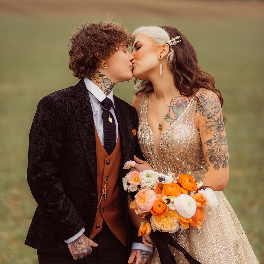 Bride in dark velvet bridal jacket with rust waistcoat and pocket square, black suit trousers and gold pocket watch standing with bride in sleeveless deep v neck sparkly gold wedding dress holding orange garden rose, white poppy, light pink carnation and foliage bridal bouquet wrapped in sparkly gold ribbon at Kingscote Barn Tetbury wedding