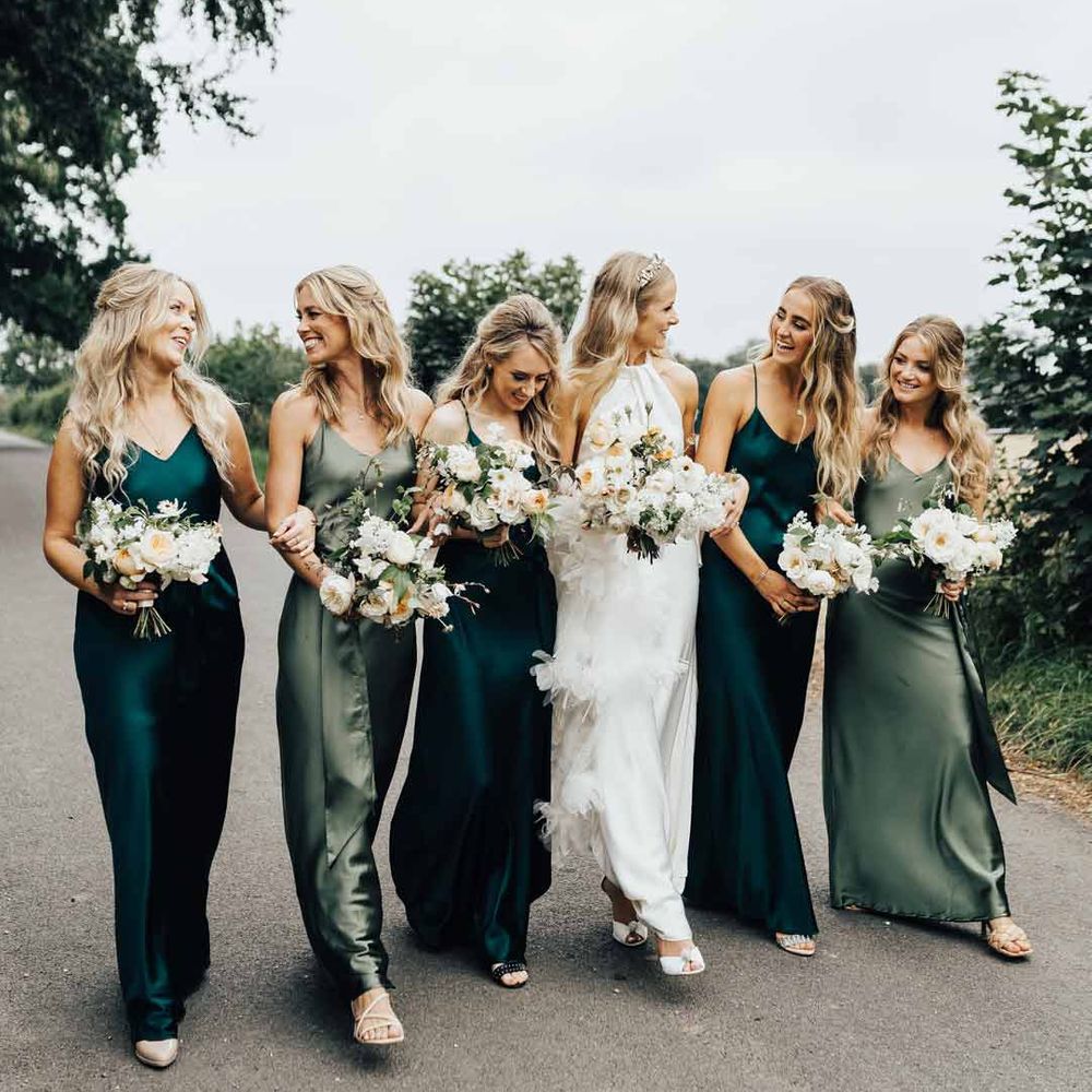 Bride in halterneck wedding dress walking with bridesmaids in mismatched green satin bridesmaid dresses 