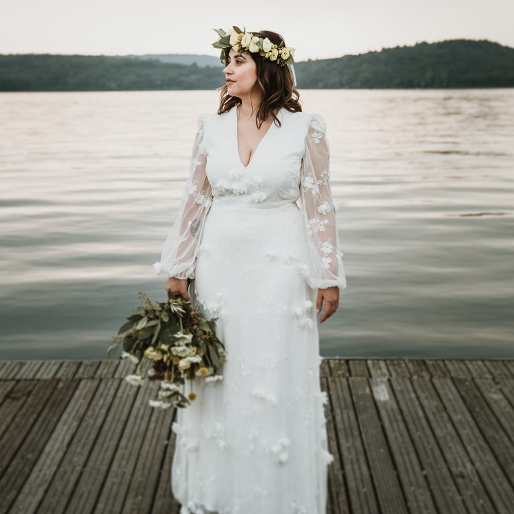Boho bride in a white wedding dress with flowers and flower crown standing by Lake Viverone in Italy