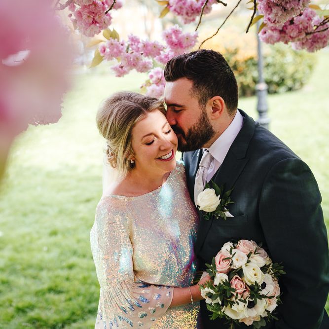 Groom kisses bride on the cheek with blossom surrounding them