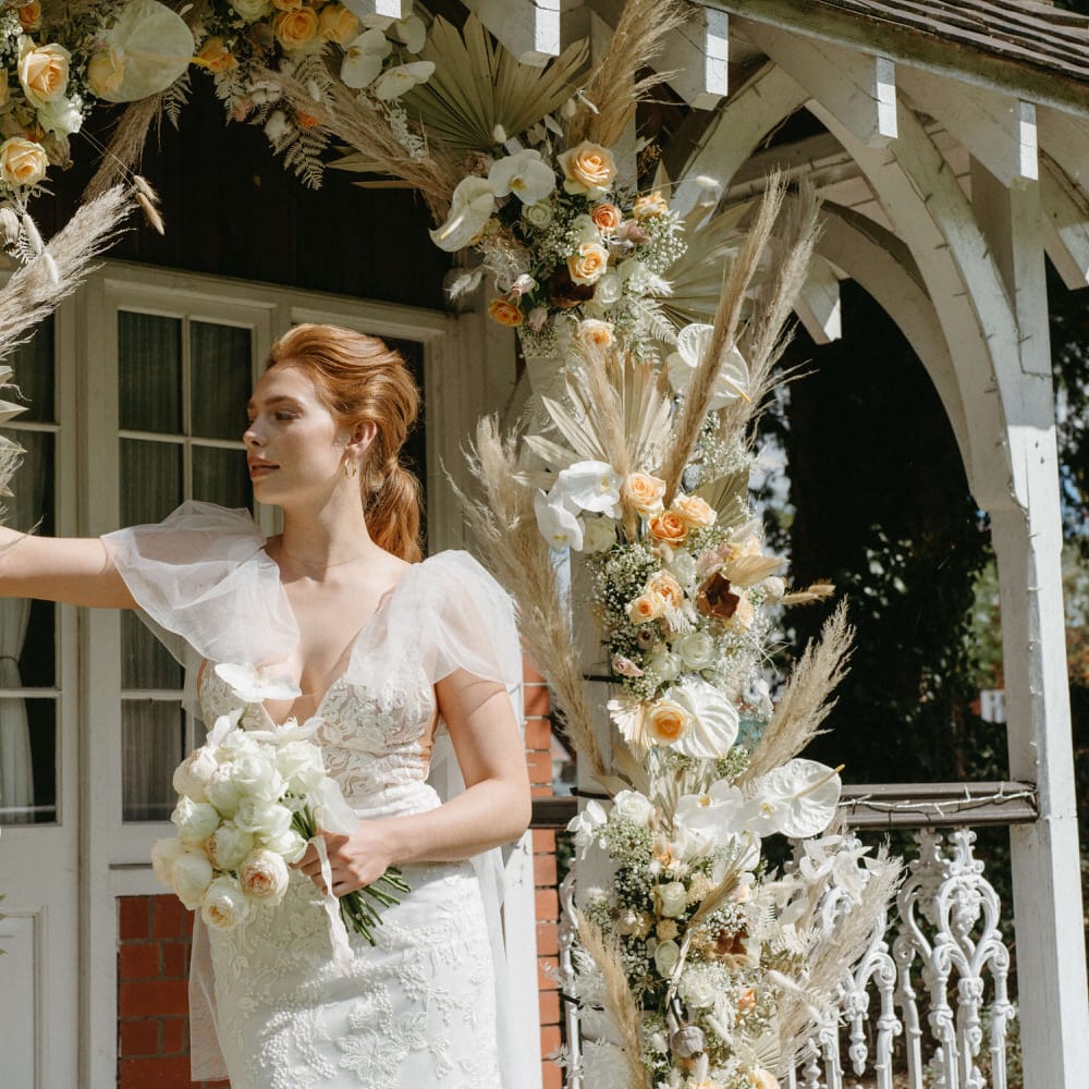 Boho bride at Berwick Lodge with puff cap sleeve wedding dress standing under an arch of dried palms, pampas grass, white anthuriums, roses and orchids