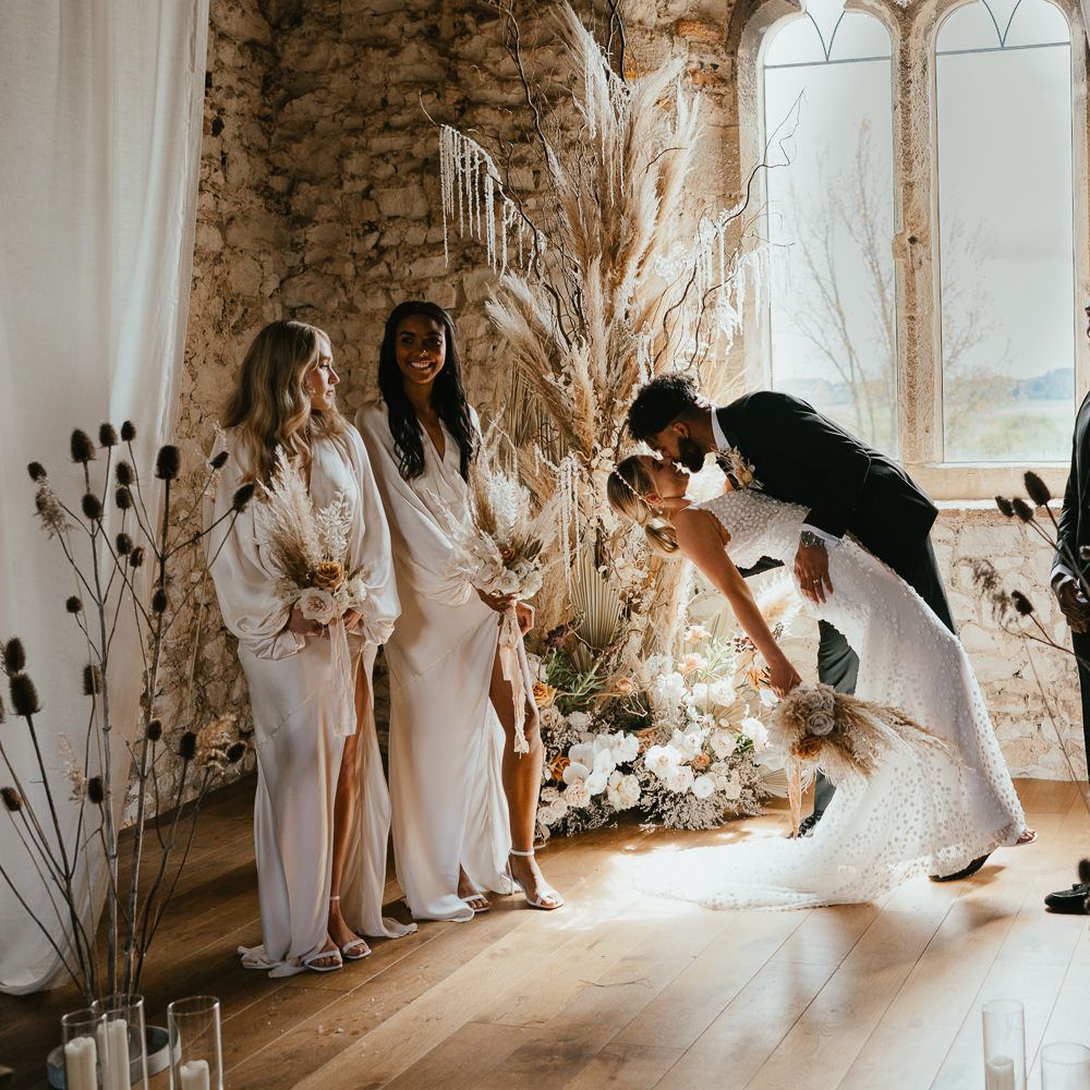 Groom leaning his bride down for a kiss at the wedding ceremony altar surrounded by fresh and dried flower arrangements and drapes. 