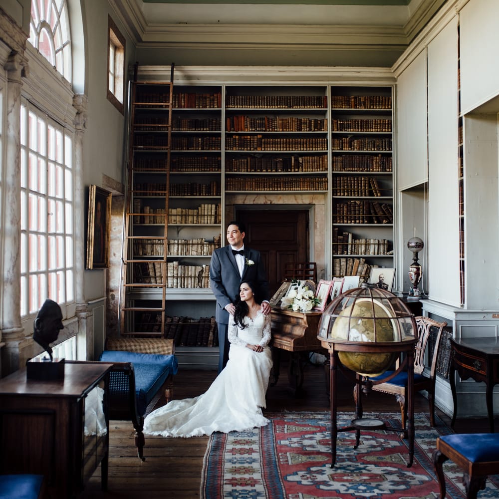 Bride sits down as groom places his hands on her shoulders within regal room filled with books 
