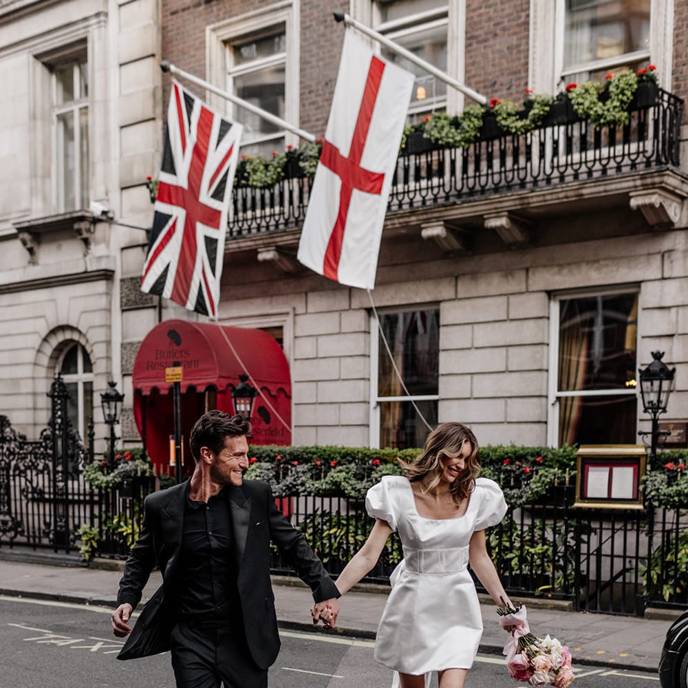 Bride and groom crossing the road in Mayfair, London in an all black suit, and short wedding dress with trailing bow detail 