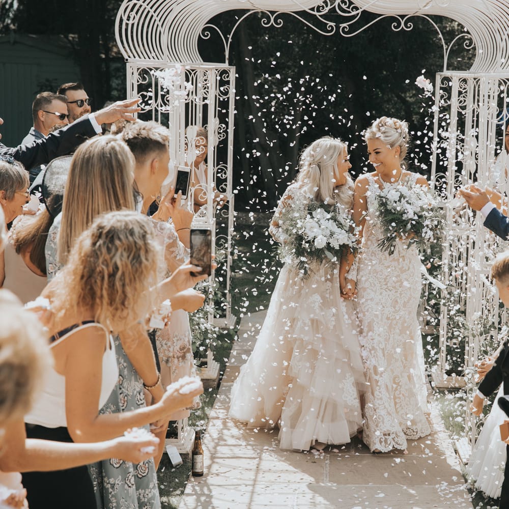 A lesbian couple walk up the aisle after their wedding ceremony through white confetti.