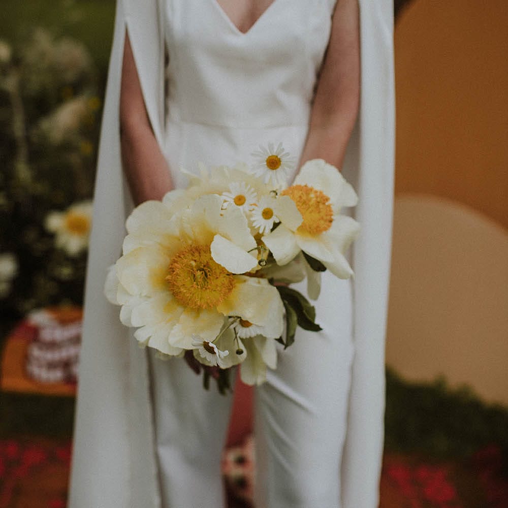 Bride in a jumpsuit and bridal cape holding a white peony and giant daisy wedding bouquet 