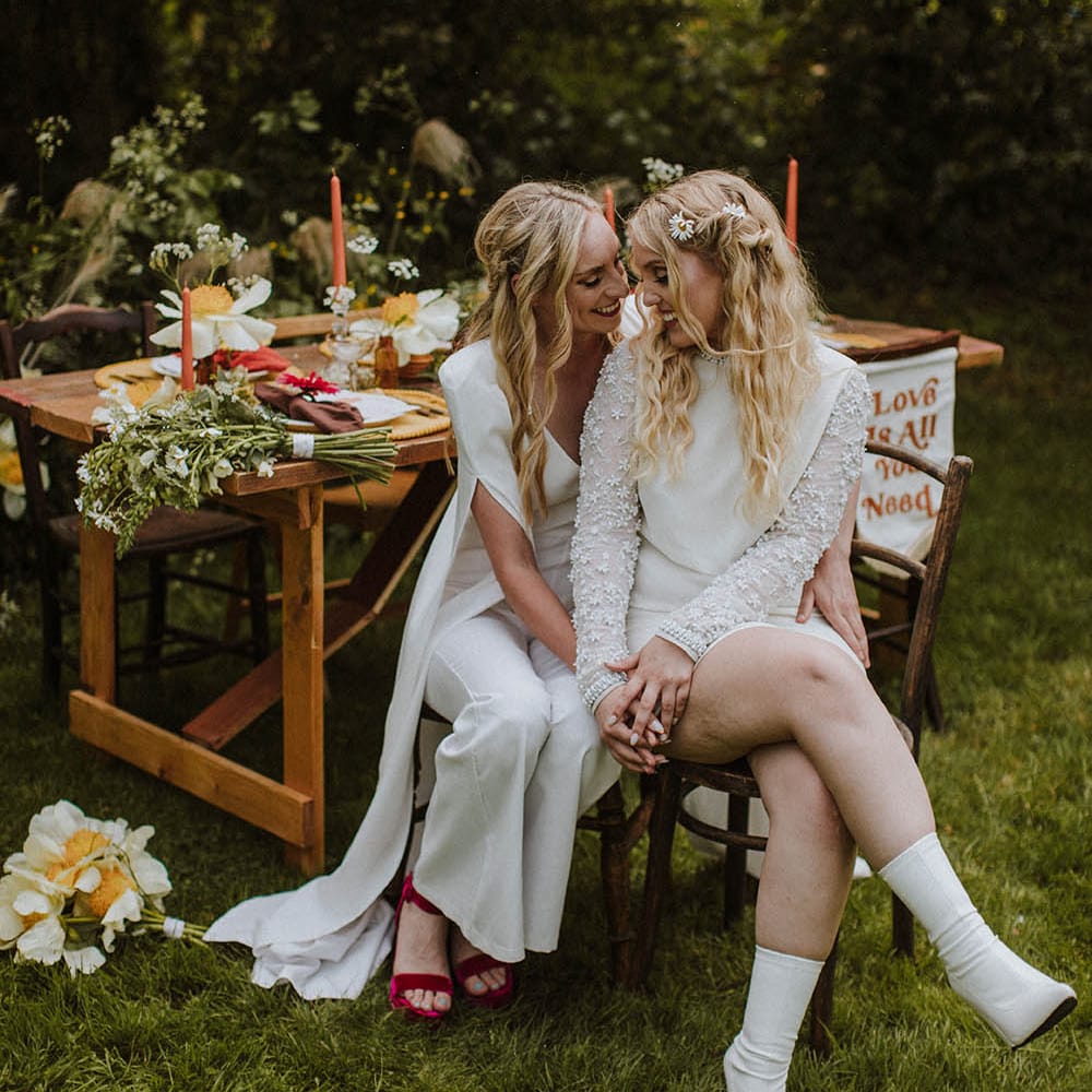 two brides with half up half down wedding hairstyles sitting on a chair at their outdoor sixties wedding 