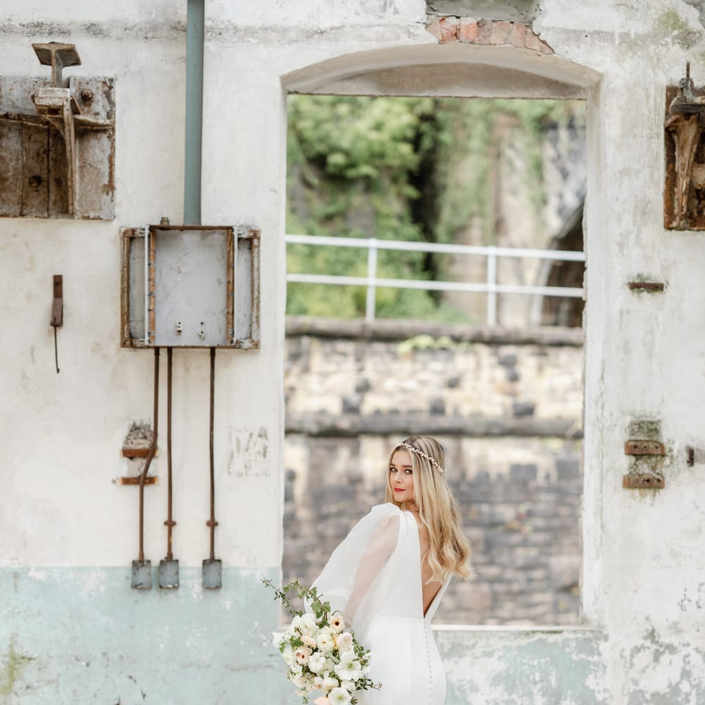 Bride in a fitted wedding dress with low V back and button detail standing in the middle of industrial wedding venue, Torr Vale Mill 