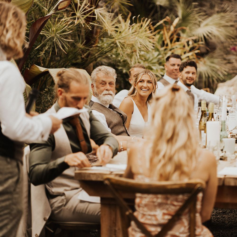 Bride smiles at groom as he makes speech in front of guests at wedding breakfast inside glasshouse at Anran Devon with wooden tables, candles and palm leaves
