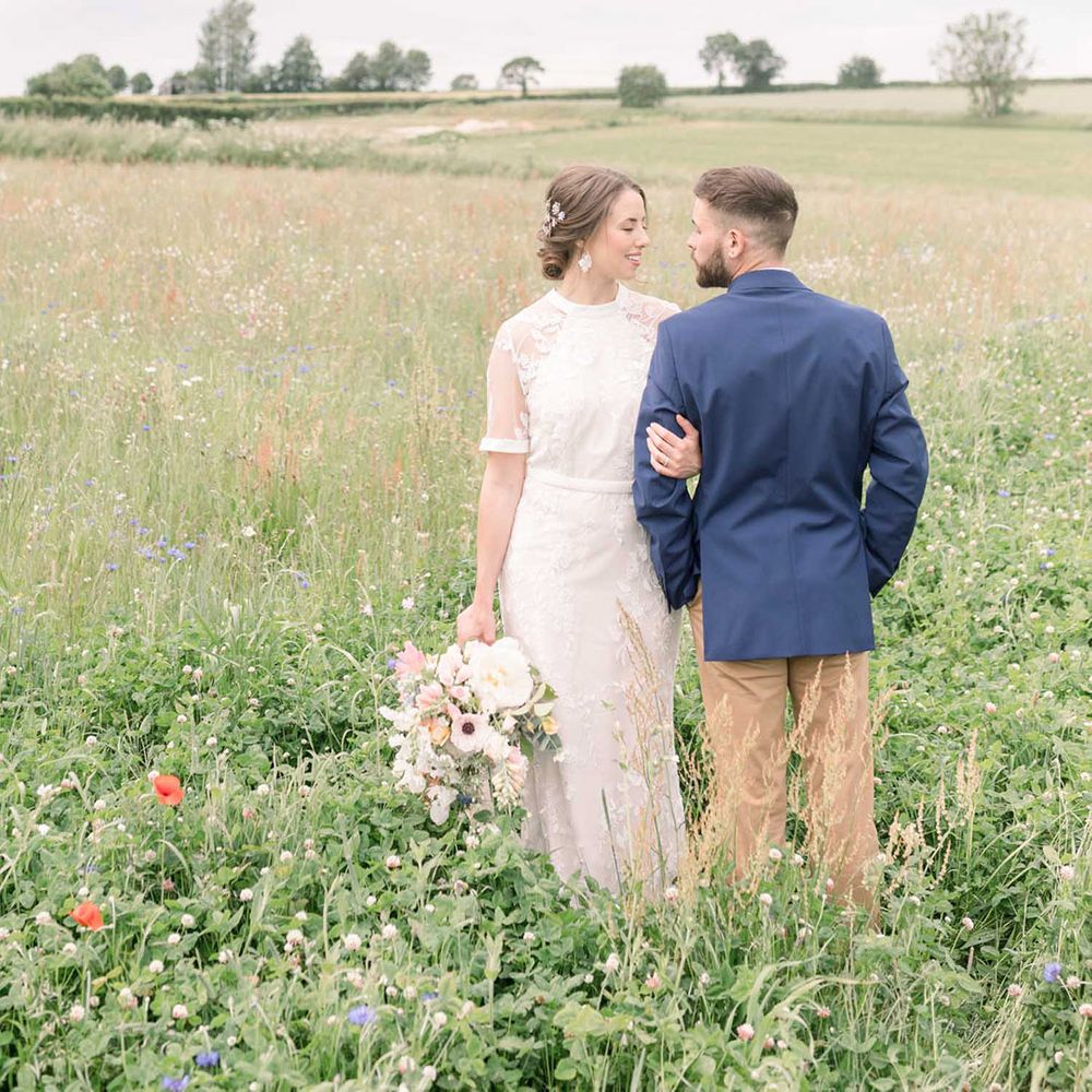 Portrait of a bride in a lace Phase Eight wedding dress holding a wildflower bouquet in a field with her groom in a navy blue blazer and chinos