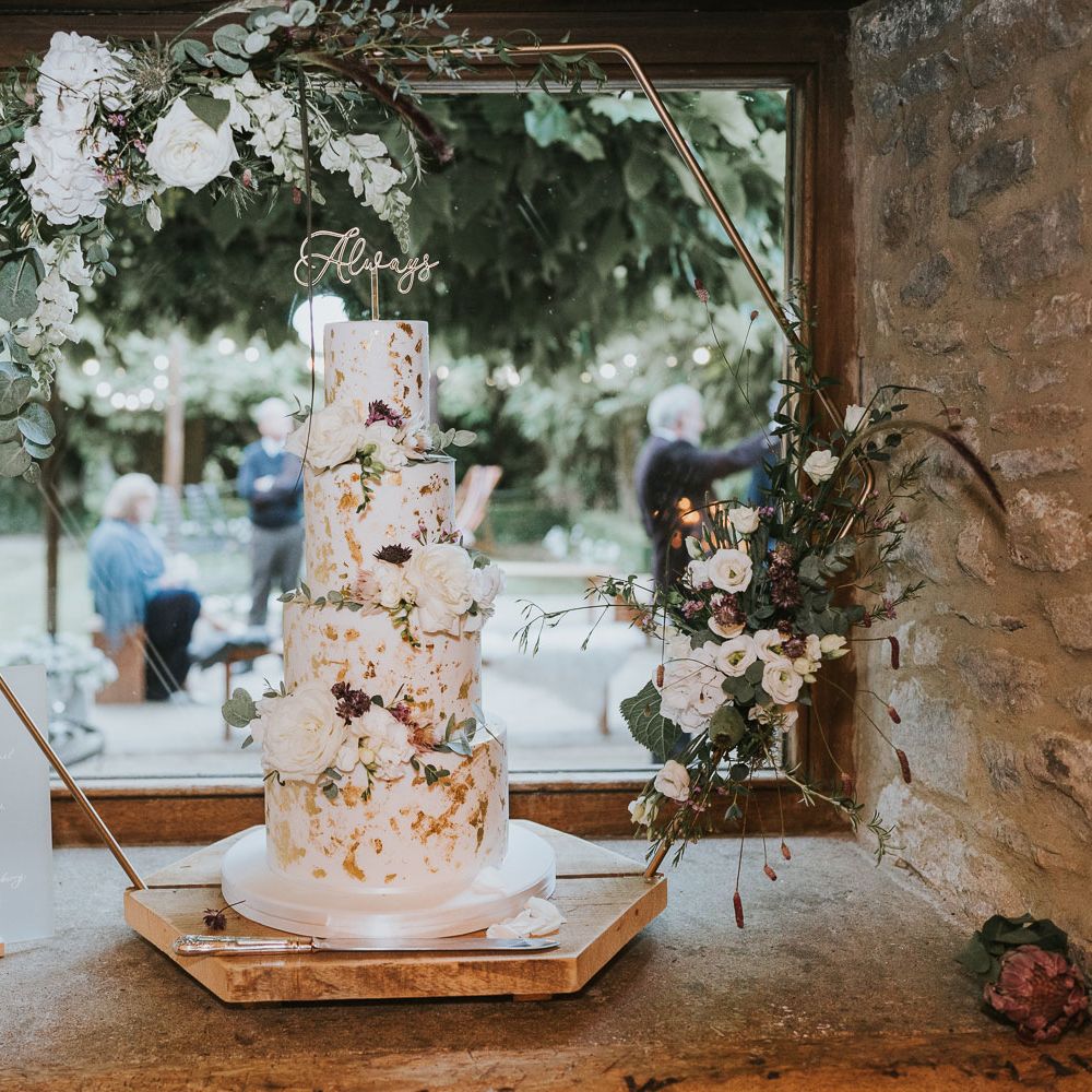 White four tier wedding cake with gold details decorated with white roses, green leaves and cake topper reading 'Always' on gold hexagonal cake stand at Tythe Barn wedding with barn wedding flowers