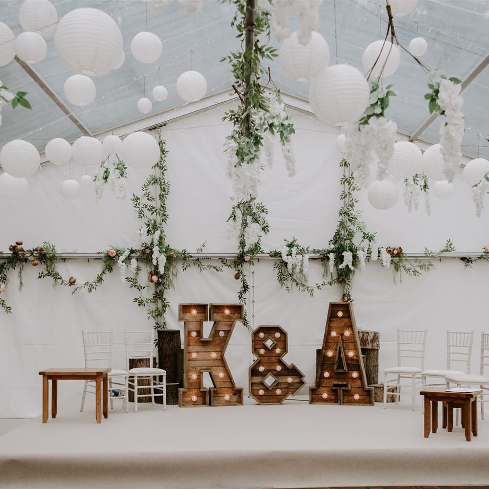 Interior of marquee wedding reception with stage, large wooden light up letters, chairs, garlands hanging from the ceiling and white paper lanterns for garden wedding in Bedfordshire