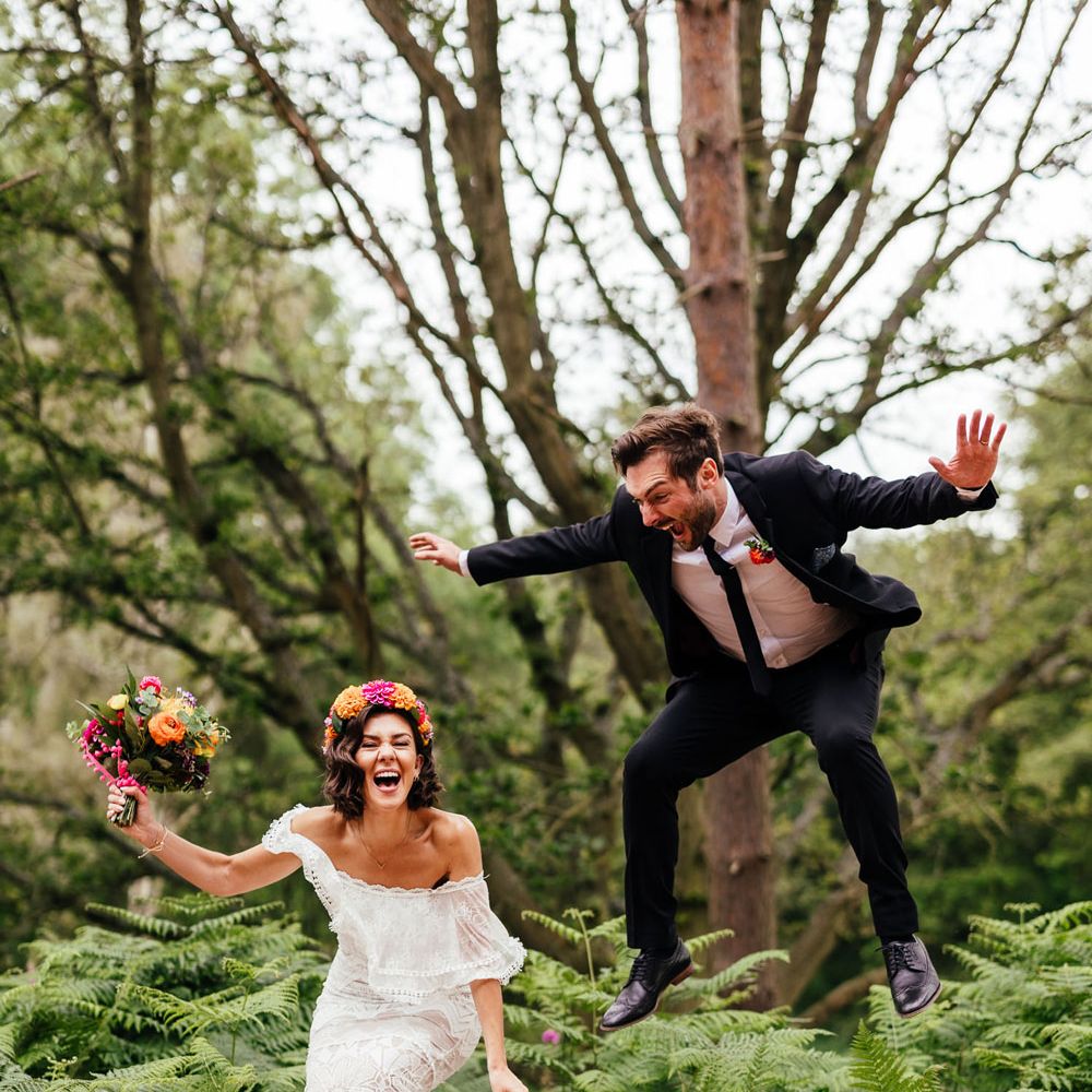 Bride in lace Grace Loves Lace wedding dress, flower crown and holding colourful bridal bouquet jumps in the woods with groom in black suit at The Bridal Barn venue