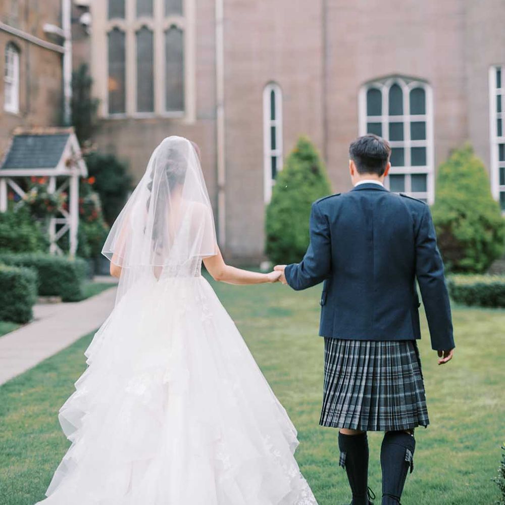 Groom in a blue tartan kilt and blazer holding hands with his bride in a princess wedding dress with layered tulle skirt at Scottish castle wedding 
