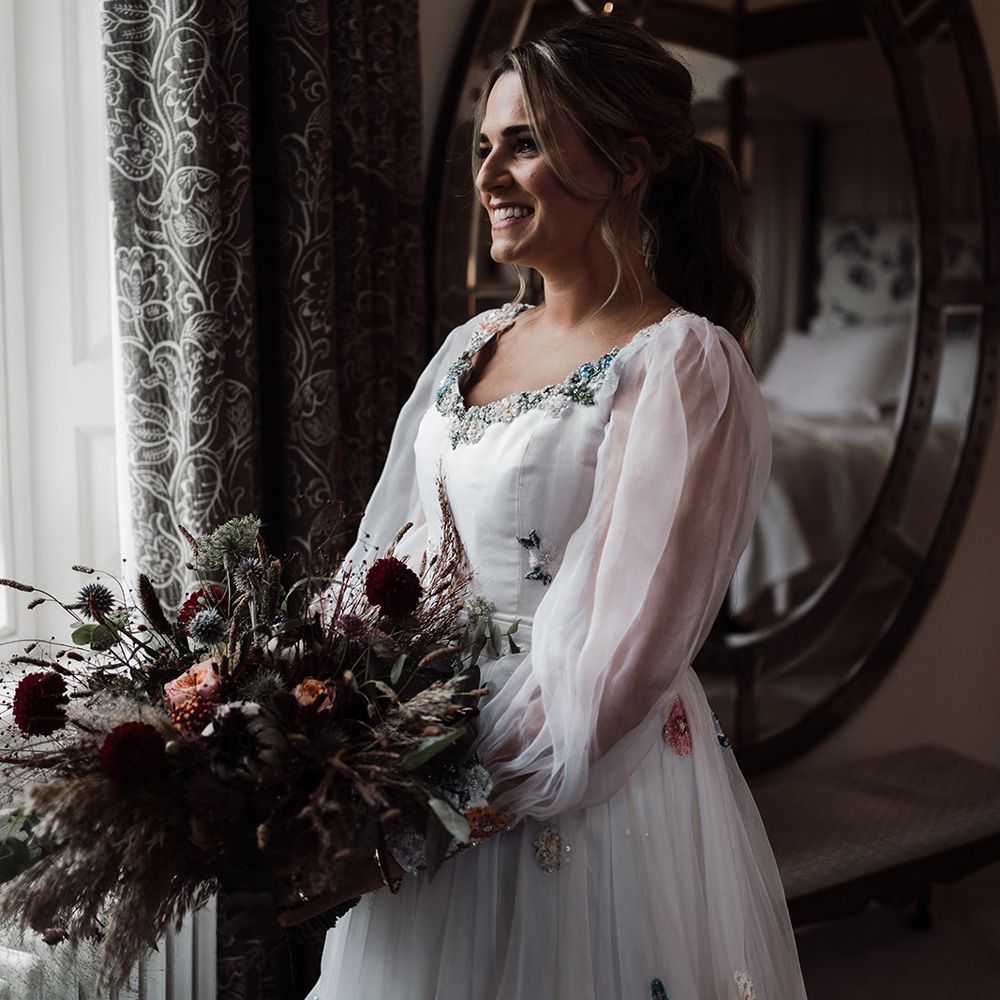 Bride wearing long mesh sleeve wedding dress and dried autumnal wedding bouquet stands by window before wedding