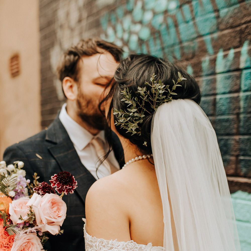 Bride in lace drop strap wedding dress with foliage hair decor and veil kisses groom by street art in Liverpool after wedding ceremony