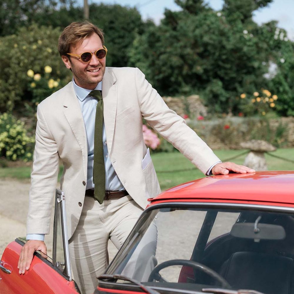 Groom in cream suit with olive green tie gets into cherry red vintage wedding car transportation