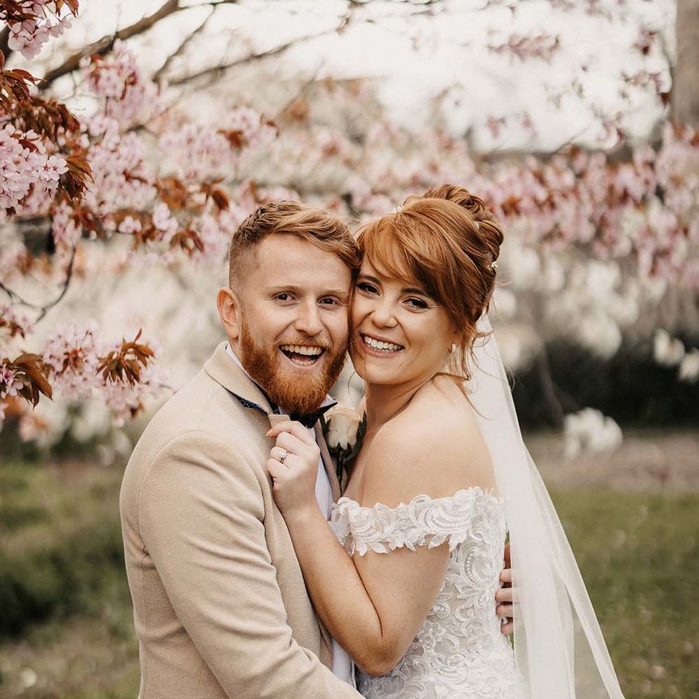 The happy newlyweds stand under pink blossom trees after their wedding ceremony in rustic barn wedding venue