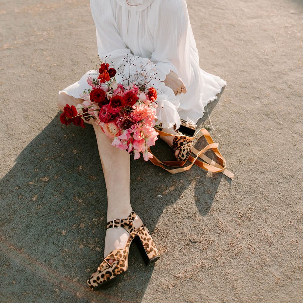 Bride in a short weddng dress with pearl encrusted birdcage veil, red lipstick and leopard print platform shoes holding a red wedding bouquet 