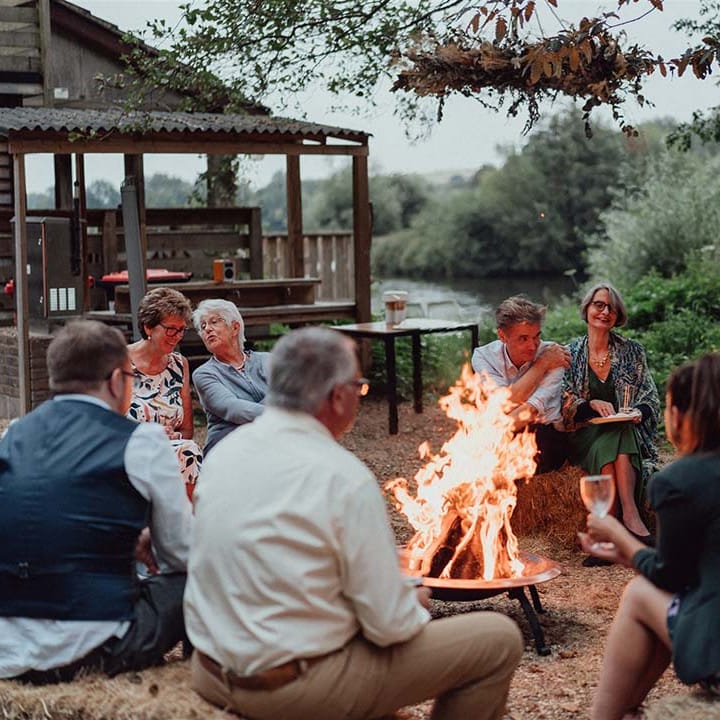 Guests sit around fire pit next to cabin near lake at wedding venue