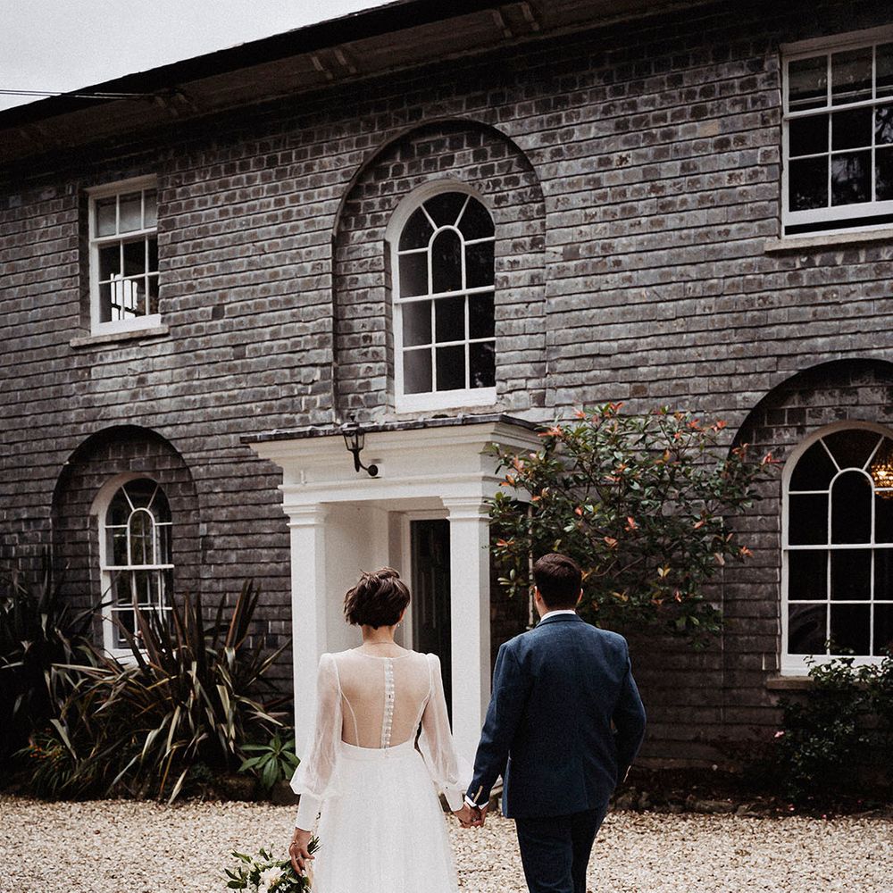Bride in sheer long sleeve wedding dress holding hands with groom in blue suit walk toward their wedding venue, Treseren