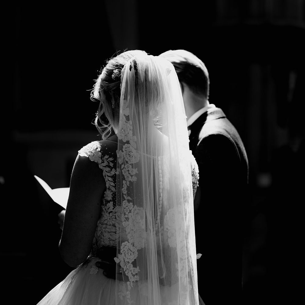 Black and white shot of the bride and groom looking through their order of service card with the bride in an illusion button back wedding dress