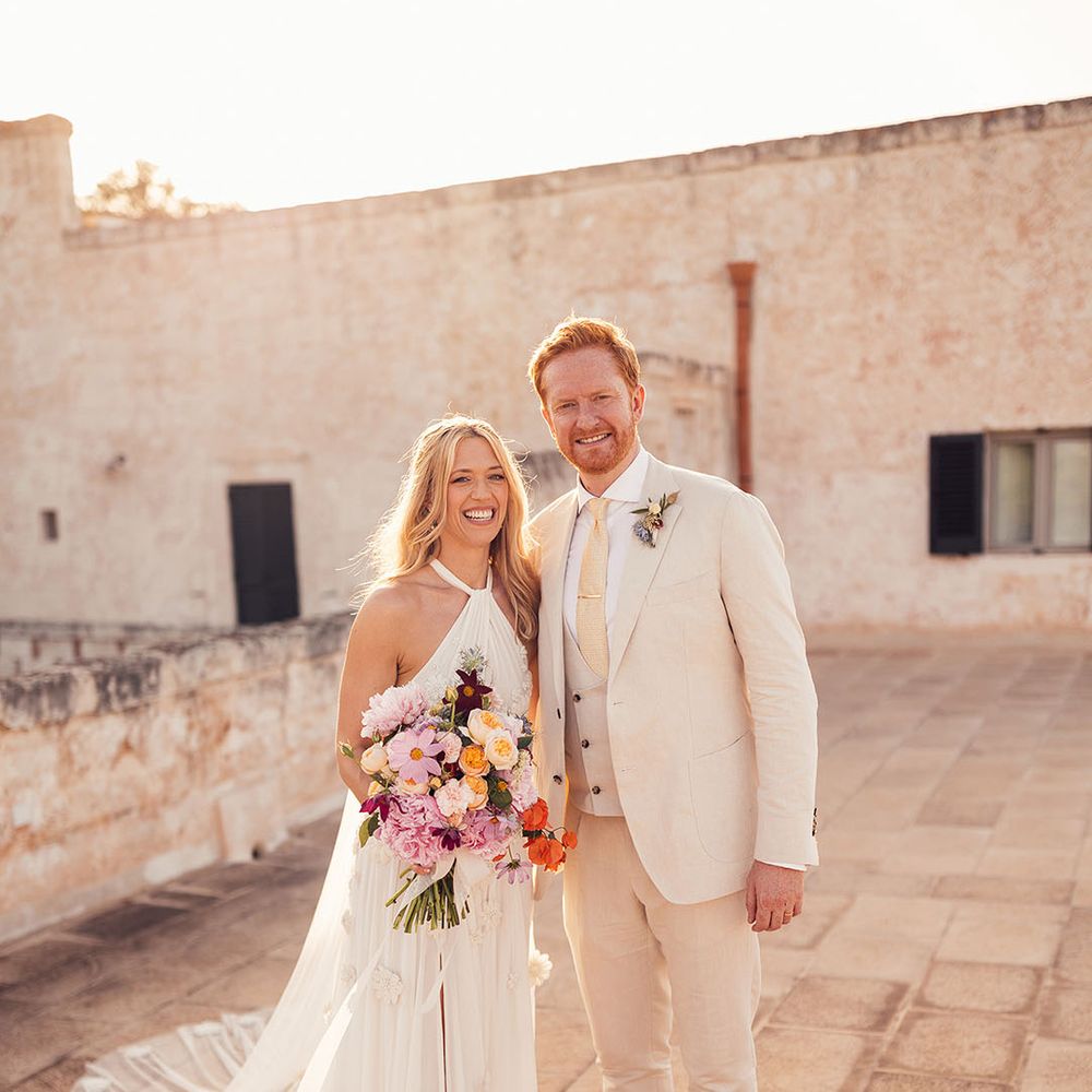 Bride holding a purple, red and pink wedding bouquet stands smiling with the groom in a cream suit 