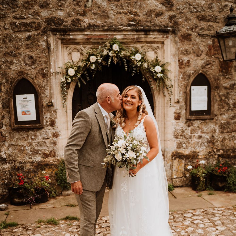 Father of the bride in a dark grey tweed suit kisses the bride on the cheek outside of the church 