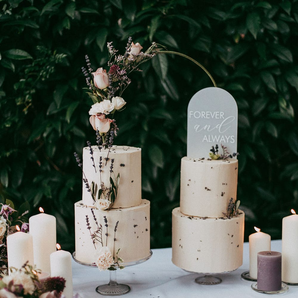Lavender and white pillar candles standing by two tiered buttercream wedding cakes with white rose and dried lavender decorations and transparent engraved cake topper and dark green half hoop
