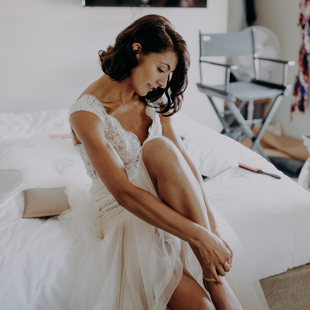 Bride in lace and tulle dress adjust the strap on her gold glittery shoes whilst sitting on bed