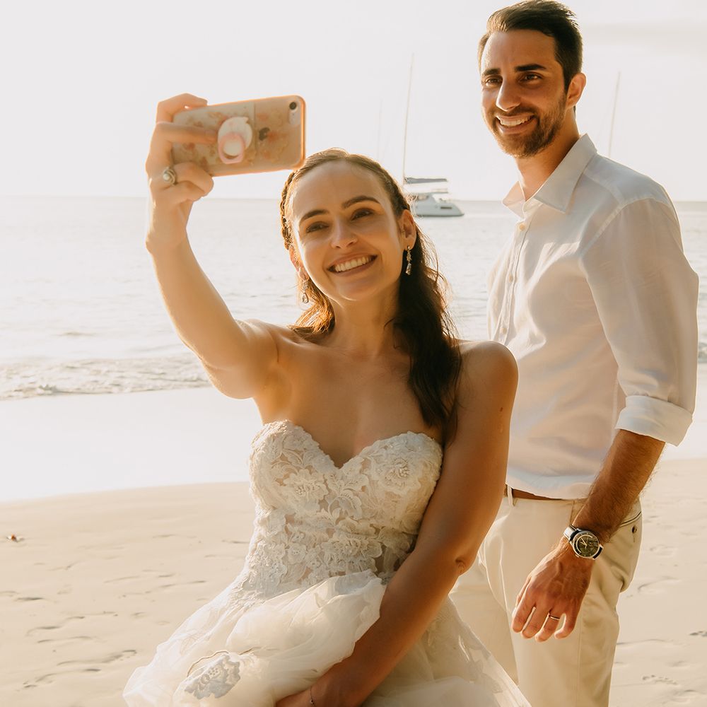 Groom in white shirt and light coloured chinos and bride in off the shoulder wedding dress with lace floral details taking a selfie on beach in Seychelles