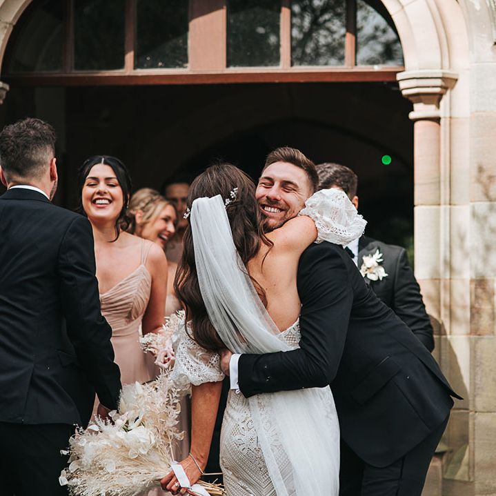 Bride in fitted boho lace wedding dress hugs a groomsman in a black tuxedo 