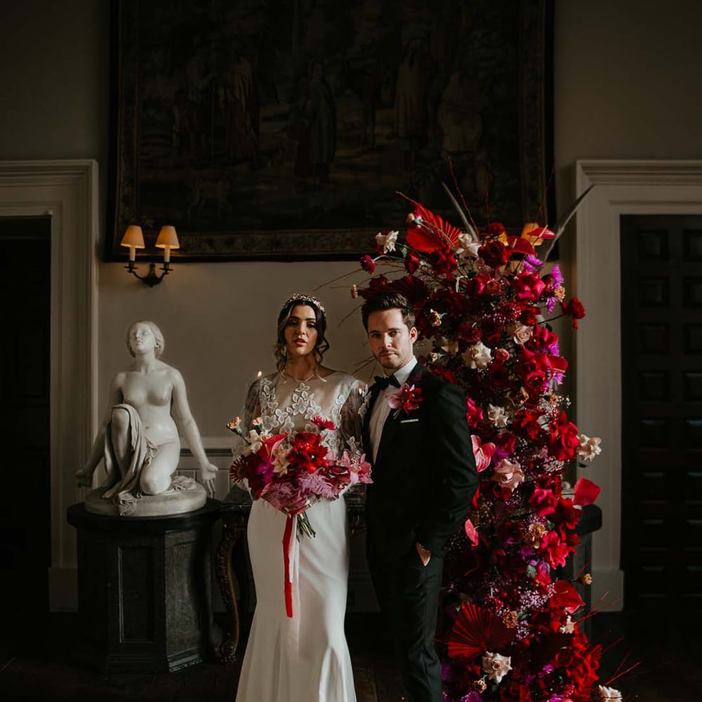 Groom in classic black tuxedo with magenta amaryllis and orchid boutonniere standing with bride in two piece bridal separates with puff sleeve sheer crop top with 3d appliqué flowers and full bodied silk skirt and gold bridal flower crown 