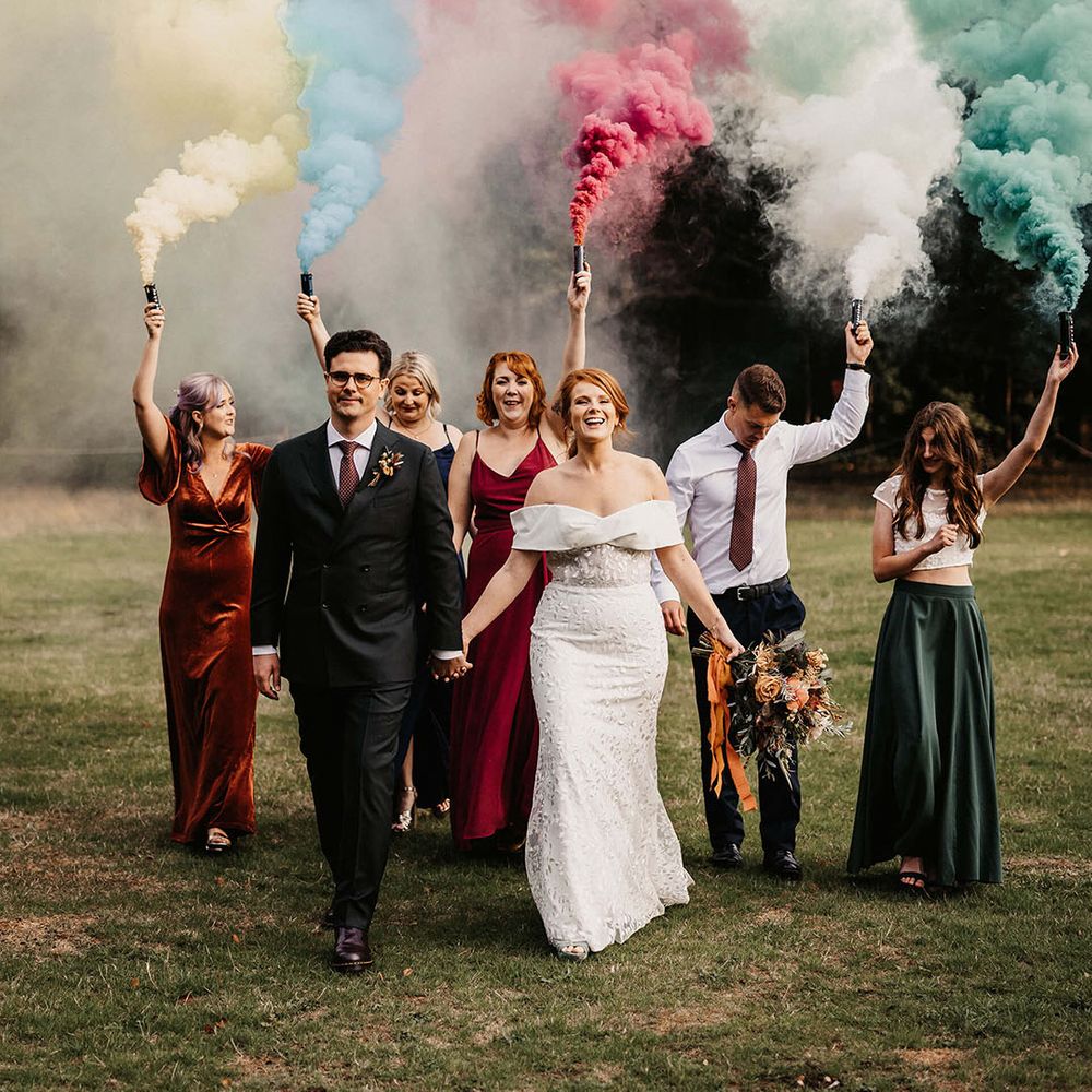 Bride & groom walk alongside wedding party holding colourful smoke bombs 