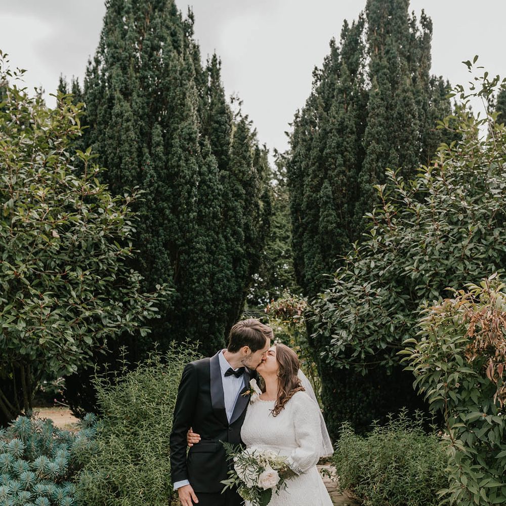 The groom in a black tuxedo smiles with the bride in a short wedding dress with bow and long sleeves at Hayne House wedding venue 