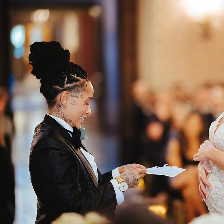 Bride in black snakeskin print tuxedo with black bowtie and white rose boutonniere reading their vows to bride in lace sleeveless v neck Enzoani wedding dress with tulle finish and church length veil holding her baby bump